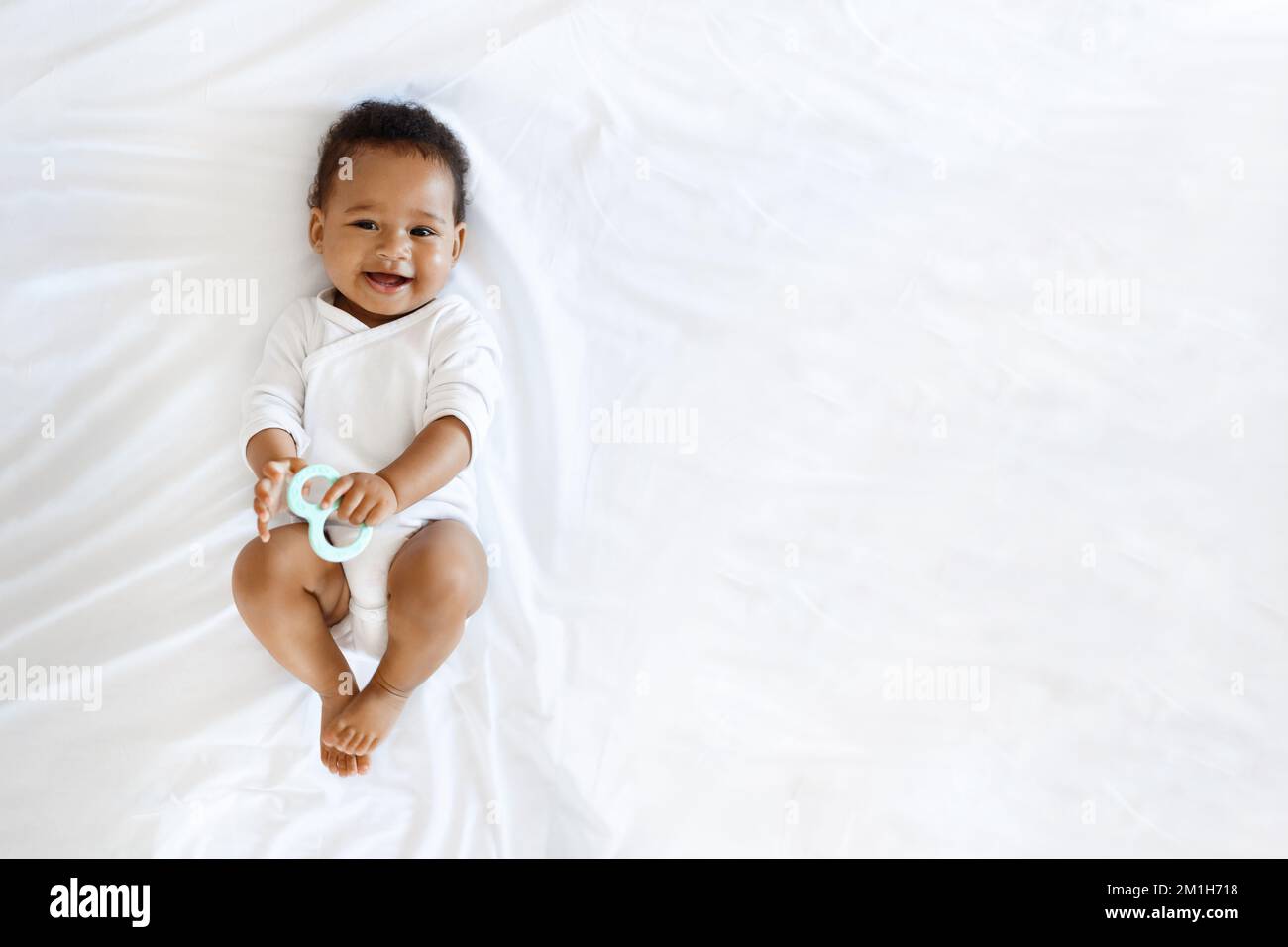 Childcare. Adorable Little Black Baby Lying On Bed With Teether In Hand Stock Photo