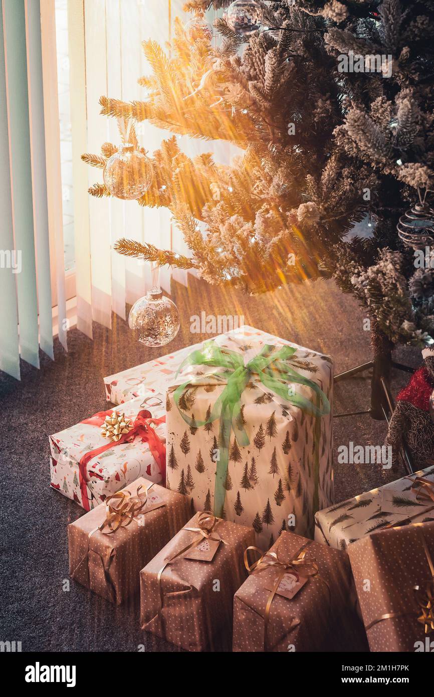 Presents wrapped under a Christmas tree as sun rays shine through vertical blinds on patio doors next to the tree. Stock Photo