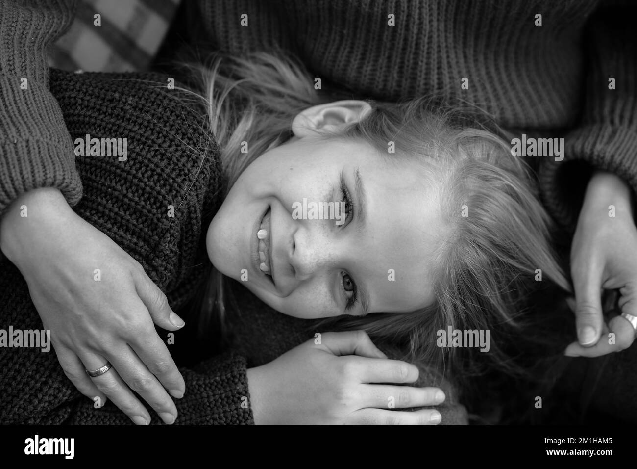 Black and white portrait of cheerful wonderful teenage girl child with long hair wearing sweater lying on legs of woman. Stock Photo