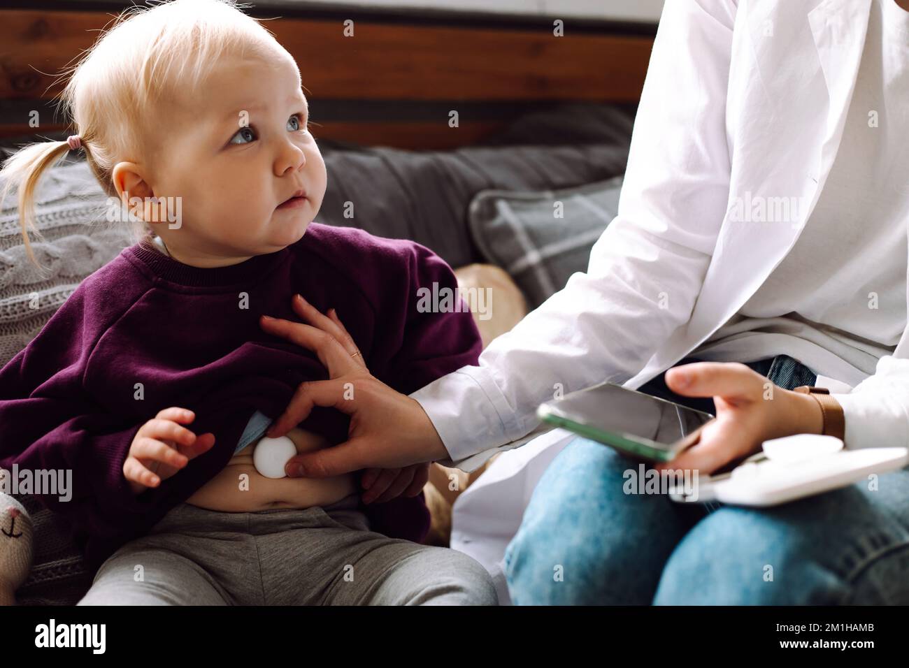 Sad little girl sitting on sofa, looking at primary care pediatrician measuring temperature with digital thermometer. Stock Photo