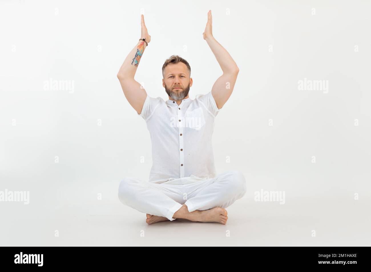Concentrated sporty middle-aged bearded man sitting with crossed legs on floor, raising hands on white background. Yoga. Stock Photo