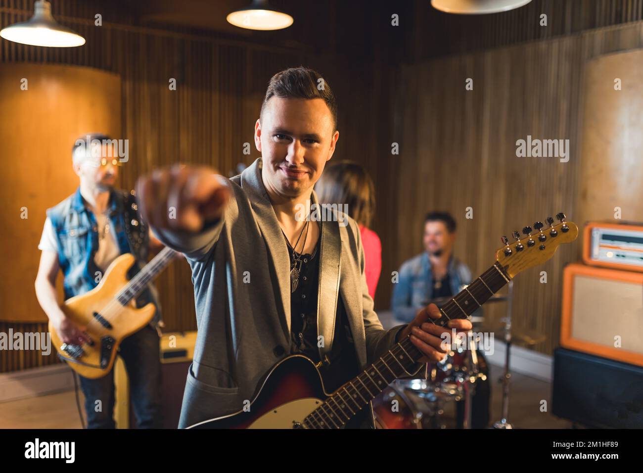 Positive caucasian male guitarist in grey jacket holding his electric guitar and pointing to the camera with index finger. Blurred hand in the foreground. Garage band. High quality photo Stock Photo