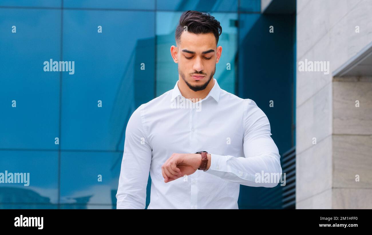 Serious upset angry hispanic indian man bearded guy disgruntled businessman boss wears formal stylish shirt stands outdoors looking at wrist watch Stock Photo