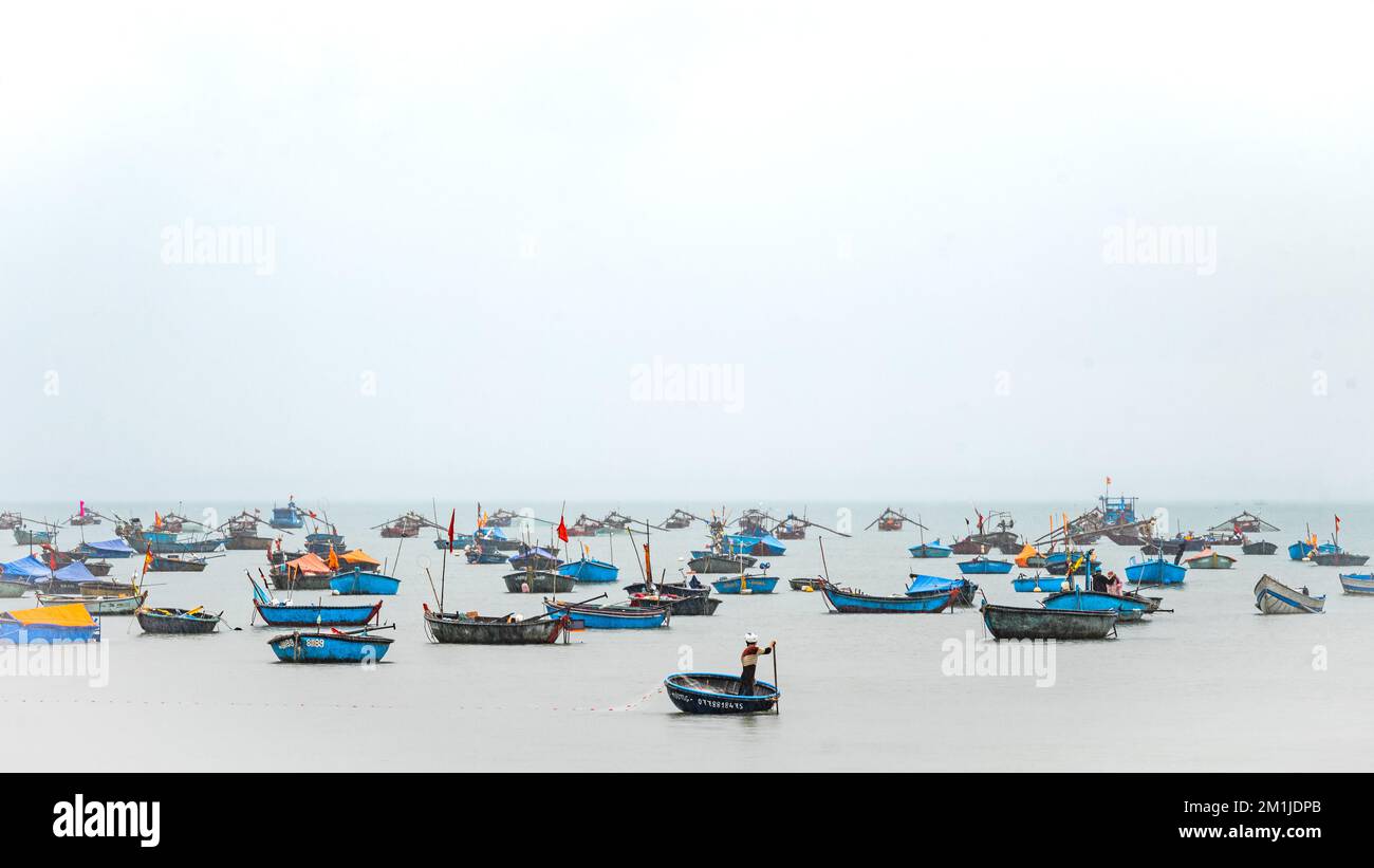 A lone Vietnamese fisherman guides his coracle as he lays his fishing net by a flotilla of moored traditional fishing boats on My Khe beach, Danang, V Stock Photo