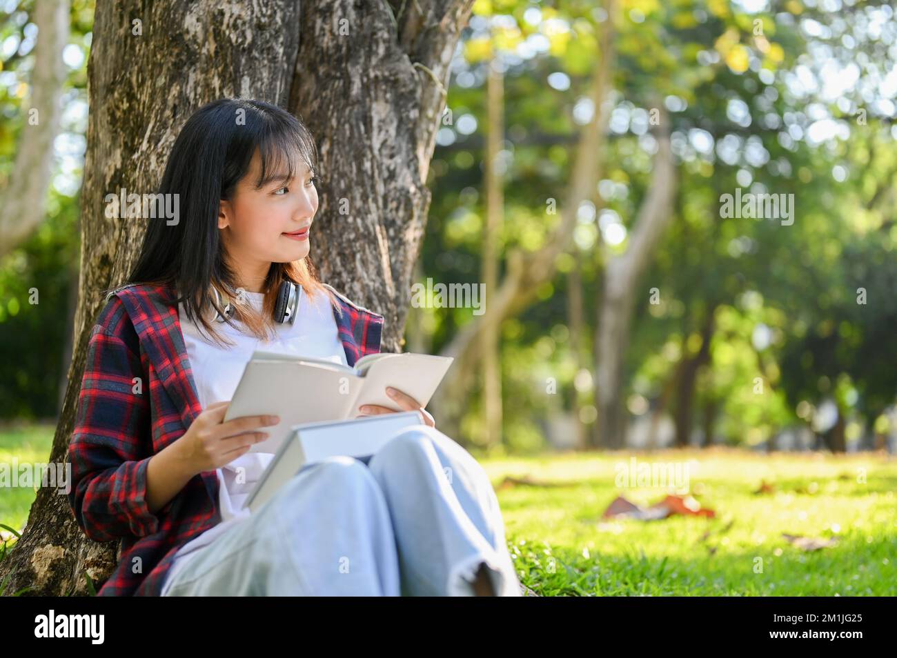 Beautiful and happy young Asian female college student reading a book, daydreaming about her success while sitting under the tree in the campus's park Stock Photo