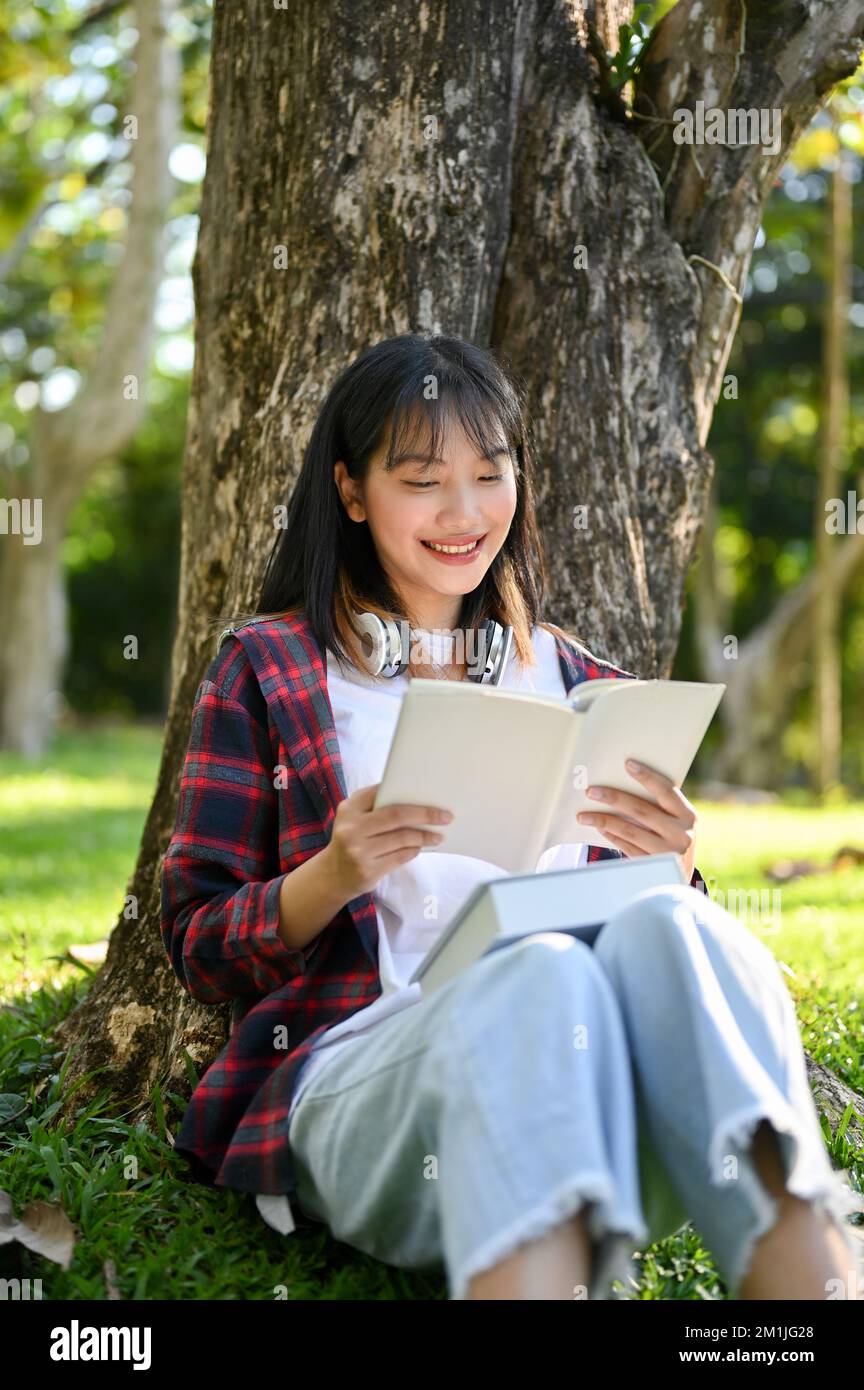 Portrait, Pretty and happy young Asian female college student reading a book while relaxes sitting under the tree in the park. Stock Photo