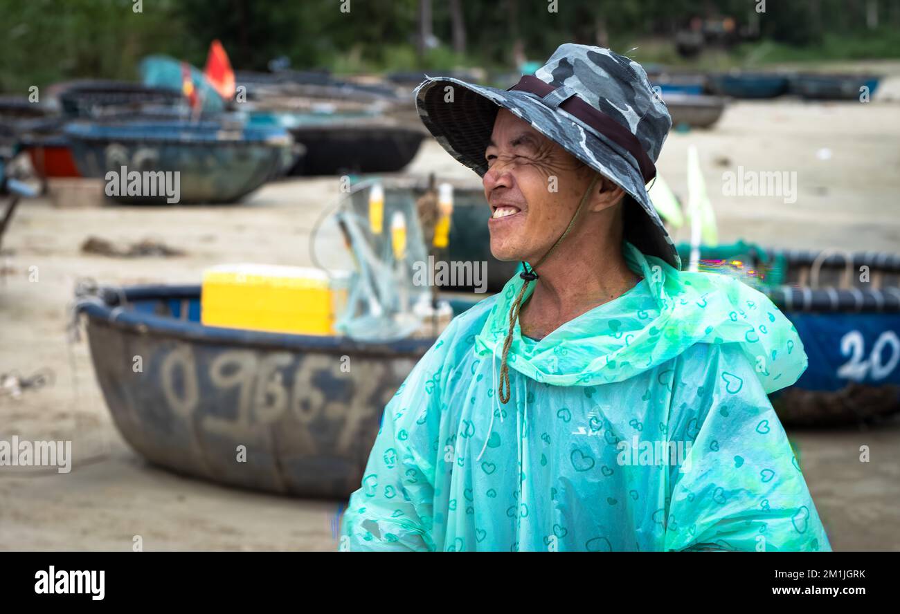 A Vietnamese fishermen wearing a raincoat with traditional coracles used for fishing on a rainy day on My Khe beach in Danang, Vietnam. Stock Photo