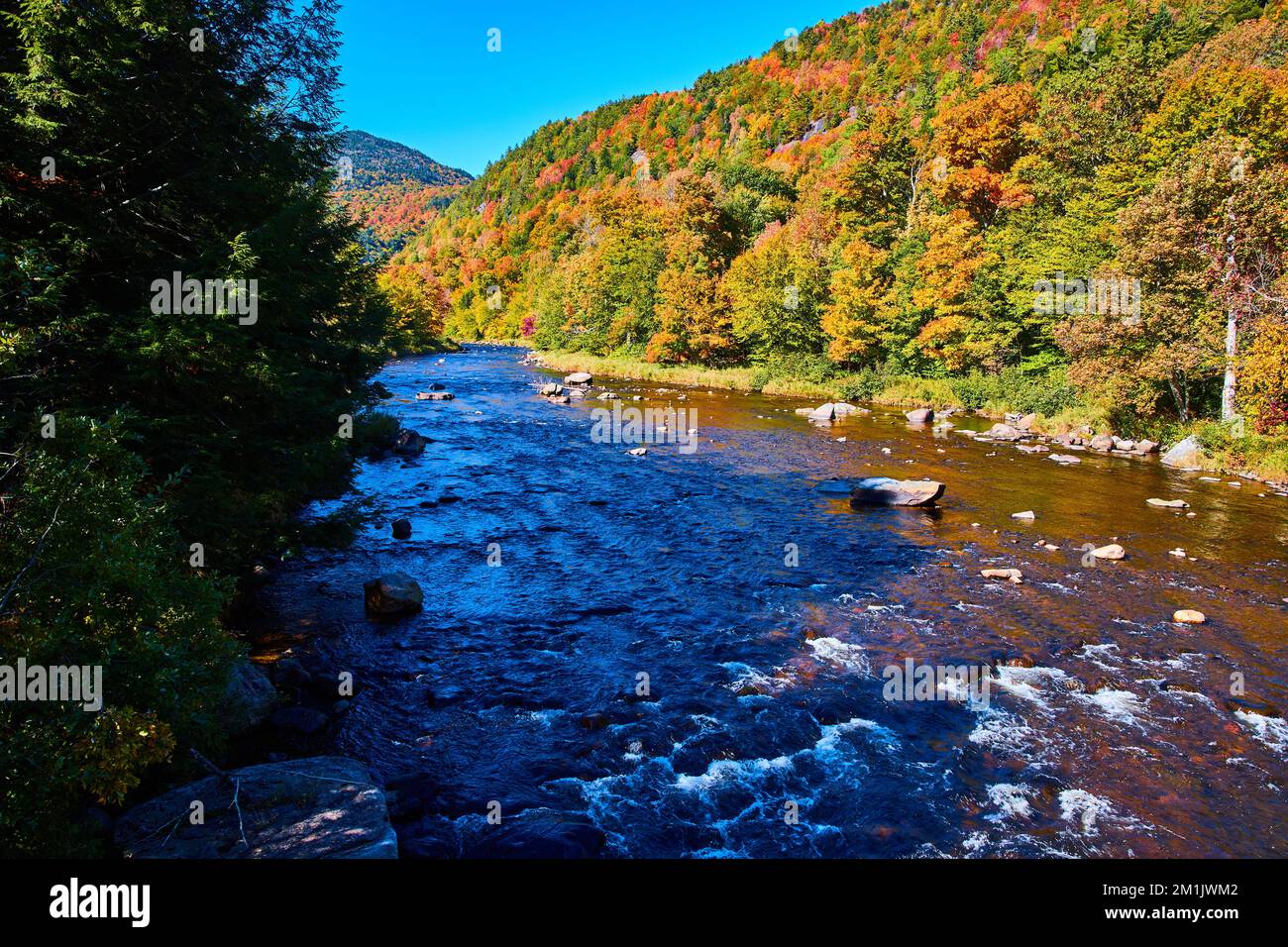 River with small rapids in shade along sunny hills of fall foliage Stock Photo