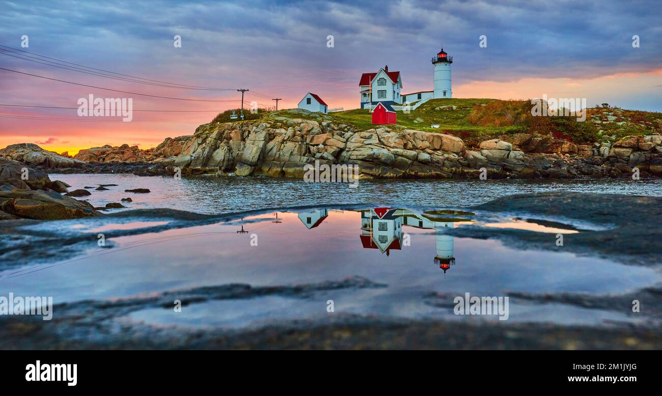 Maine lighthouse reflecting in puddle on rock during golden morning light with overcast clouds Stock Photo