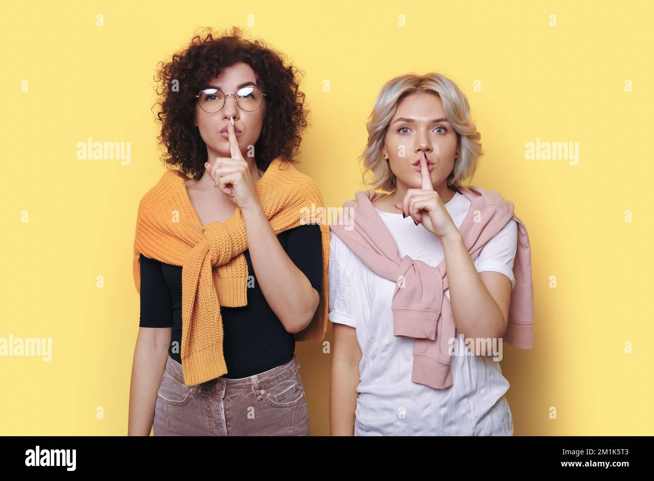 Two young women wearing casual clothes saying hush be quiet with finger on lips shhh gesture isolated on beige color background studio portrait. Stock Photo