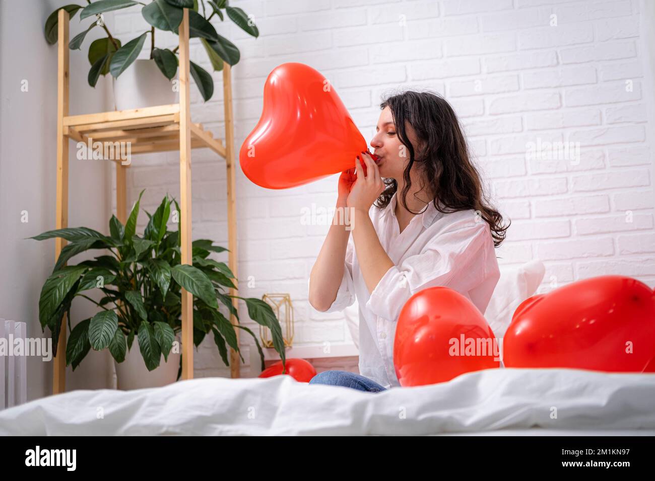 Girl is blowing air balloon at home Handsome brunette girl inflating red heart shaped balloon sitting on bed. Preparation for valentines day party Stock Photo