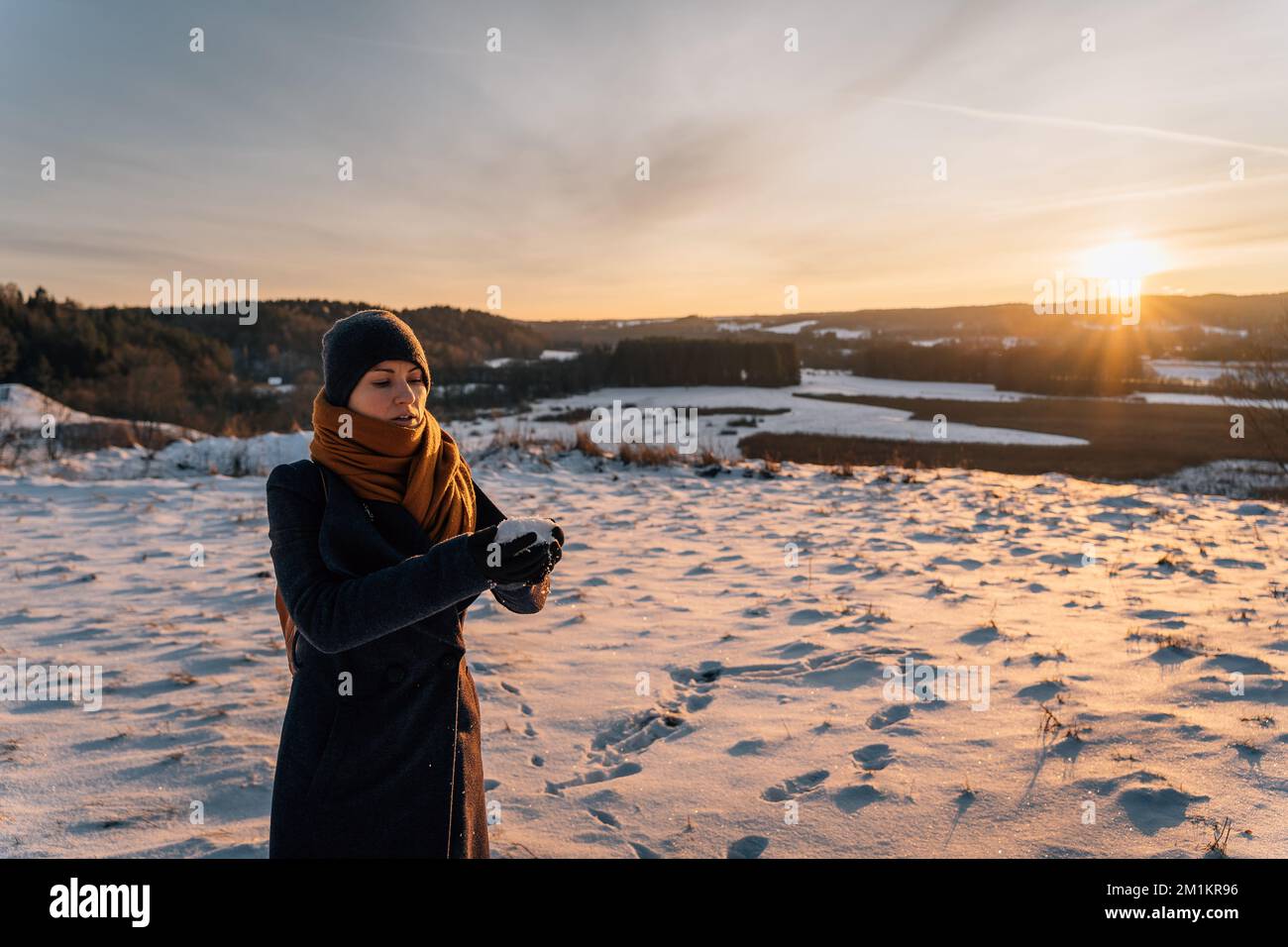 A young woman in a coat and hat at sunset stands on a snow-covered field and holds a handful of snow in her hands Stock Photo