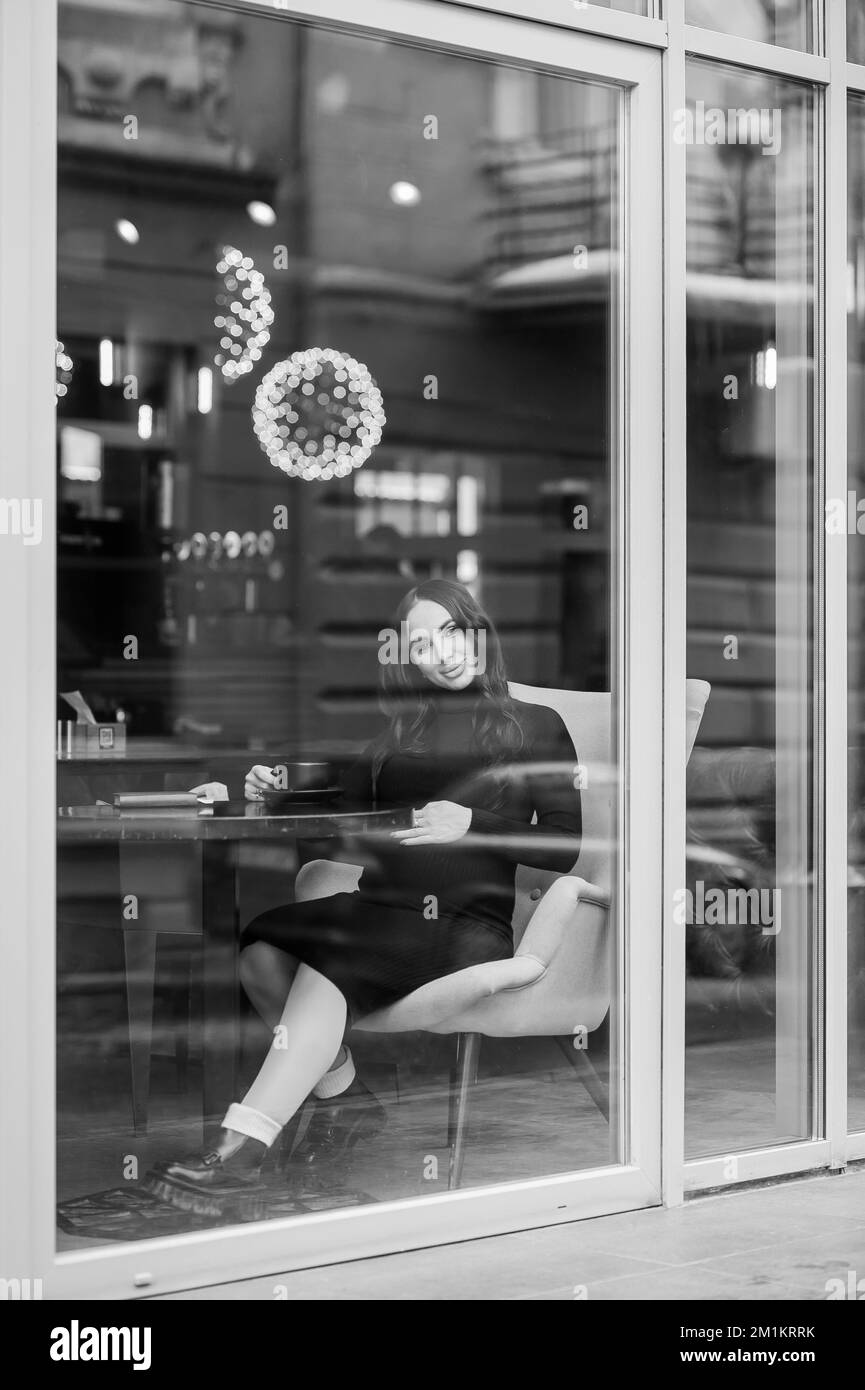 A beautiful young woman in a black dress sits in a cafe by the window ...