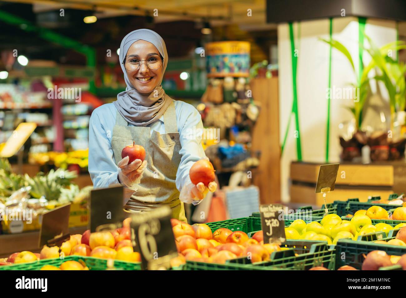 Portrait of saleswoman in supermarket, happy woman in hijab smiling and looking at camera, seller holding apples in vegetable section, Muslim woman in glasses and apron among shelves with goods. Stock Photo