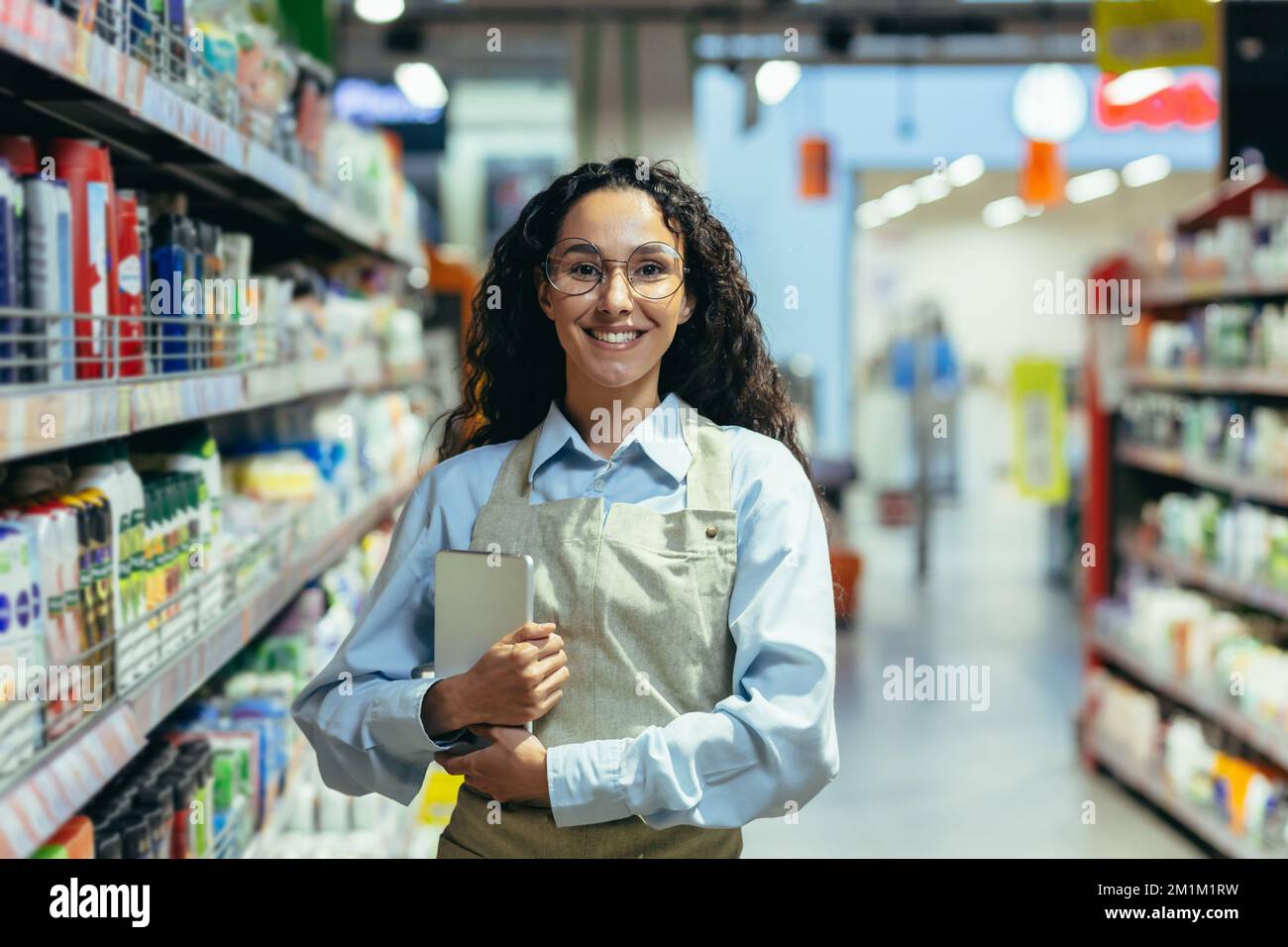 Portrait of a Hispanic female supermarket worker, saleswoman looking at the camera and smiling holding a tablet computer for product inspection, among racks and shelves in a store with goods. Stock Photo