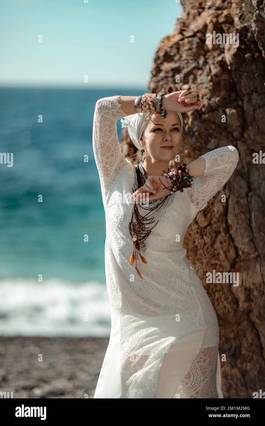Middle aged woman looks good with blond hair, boho style in white long dress on the beach decorations on her neck and arms. Stock Photo