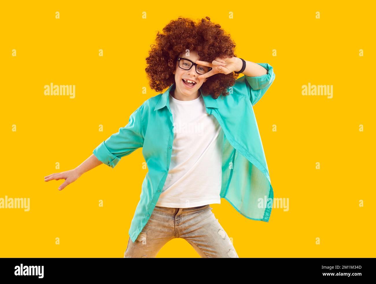Happy funny cheerful little boy in curly wig dancing on orange studio background Stock Photo