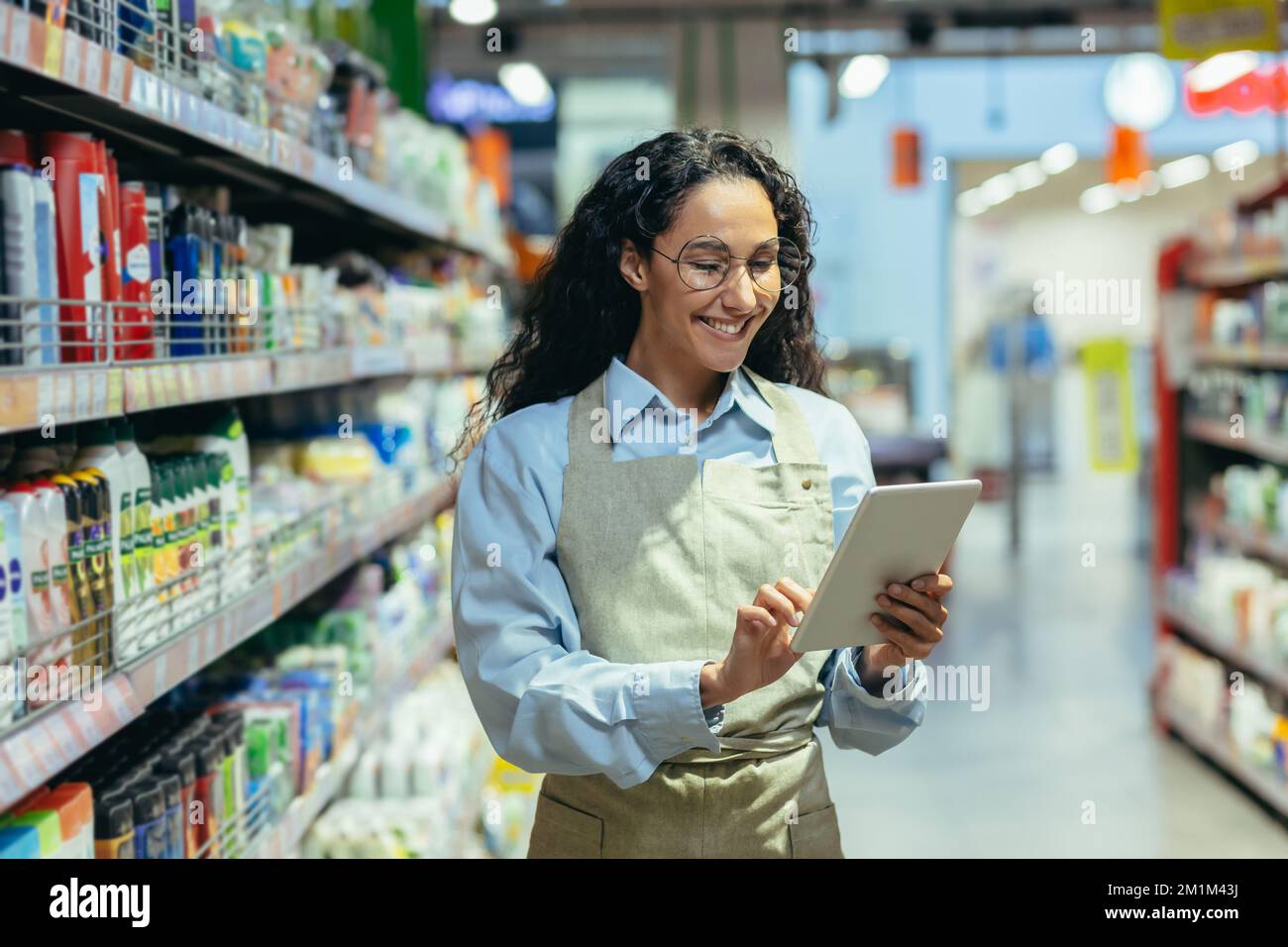 Latin American woman seller happy and smiling uses tablet computer in supermarket to check stock availability, shop assistant among shelves with goods in apron. Stock Photo