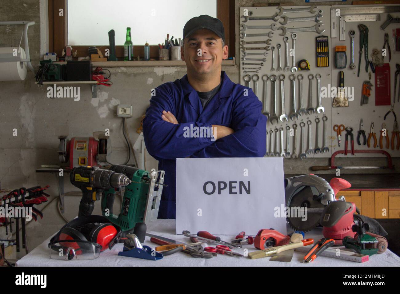 Image of a smiling handyman in his workshop with a flat bench of work tools and an open sign. Starting a new business of a craftsman Stock Photo