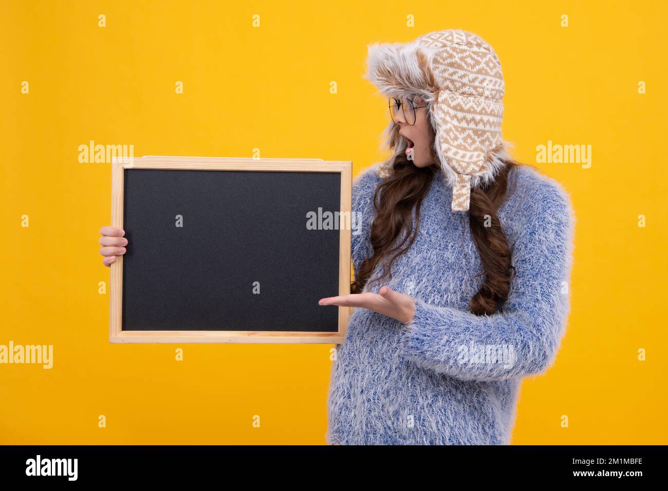 Teenage girl child in warm winter hat and sweater holding blackboard, isolated on a yellow background. Winter school sale, copy space, mockup Stock Photo