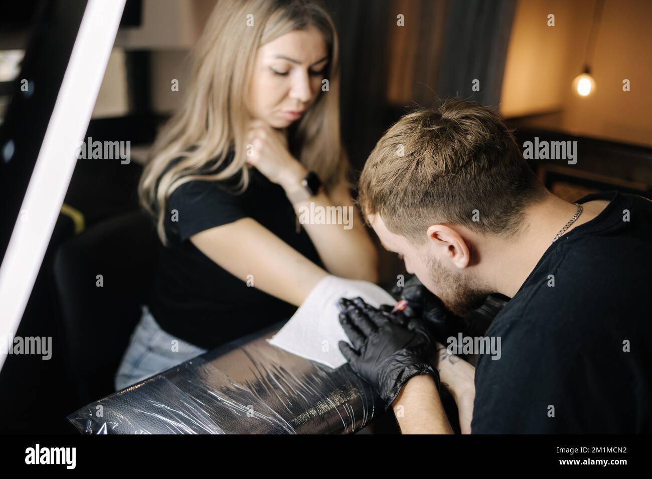 Young smiled tattoo artist is tattooing a woman's hand. Wireless tattoo machine, safety and hygiene at work. Close-up of tattoo artist work. Tattoo Stock Photo