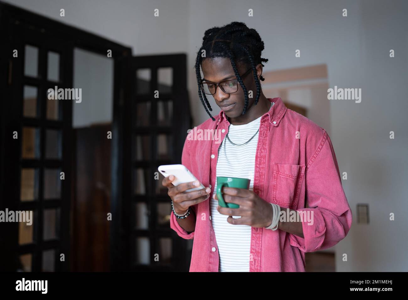 Young African man holding cup of tea and smartphone texting message while spending free time at home Stock Photo