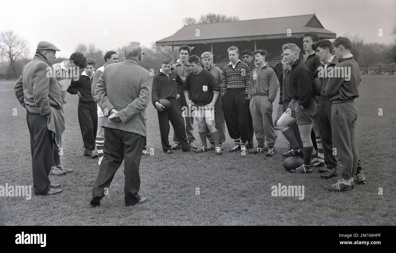 1950s, historical, members of the English Schools Rugby Football Union team at a training session outside on a pitch standing together getting instructions from one of the two adult male coaches in attendance, England, UK. All the boys are in the over 15 group, with some in shorts, but many wearing the baggy trousered cotton tracksuits of the era. Stock Photo