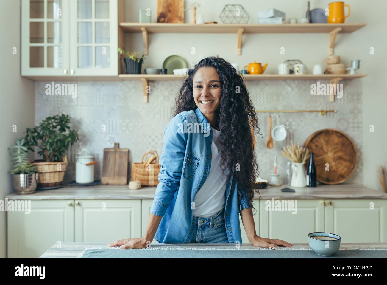 Portrait of happy hispanic woman at home in kitchen, woman with curly hair smiling and looking at camera. Stock Photo