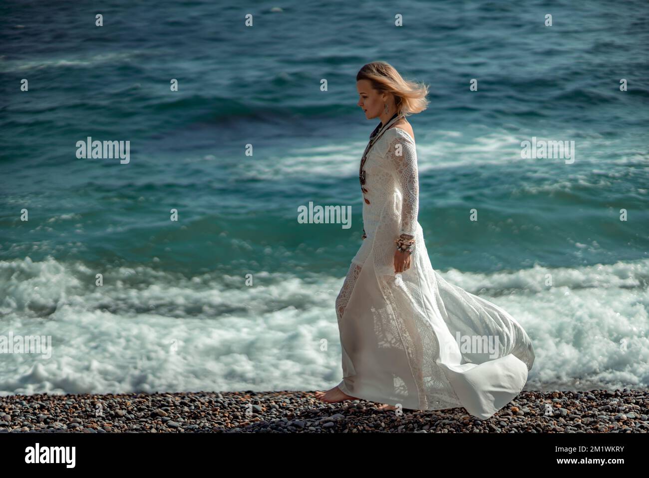 Middle aged woman looks good with blond hair, boho style in white long dress on the beach decorations on her neck and arms. Stock Photo