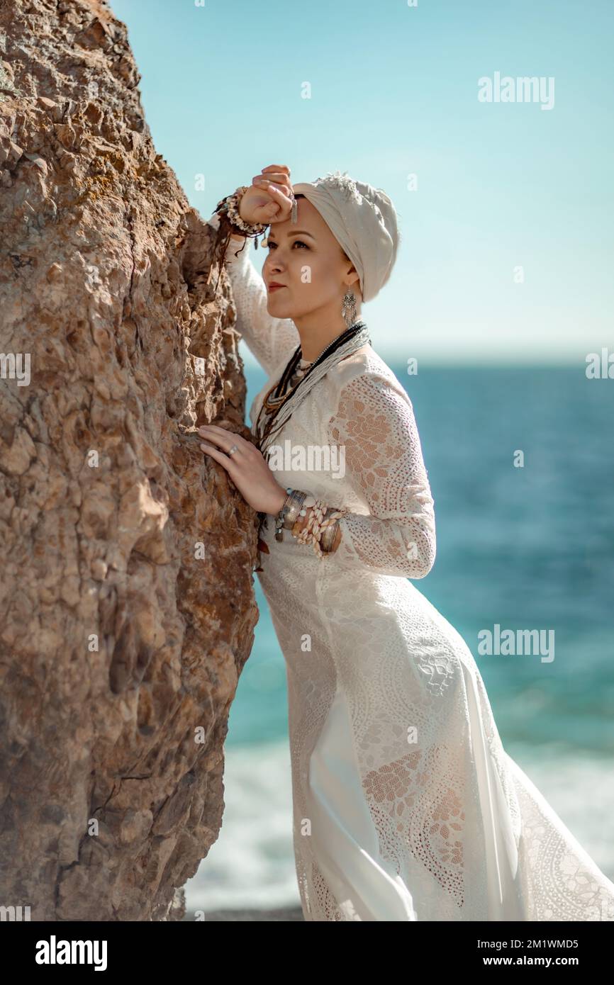 Middle aged woman looks good with blond hair, boho style in white long dress on the beach decorations on her neck and arms. Stock Photo