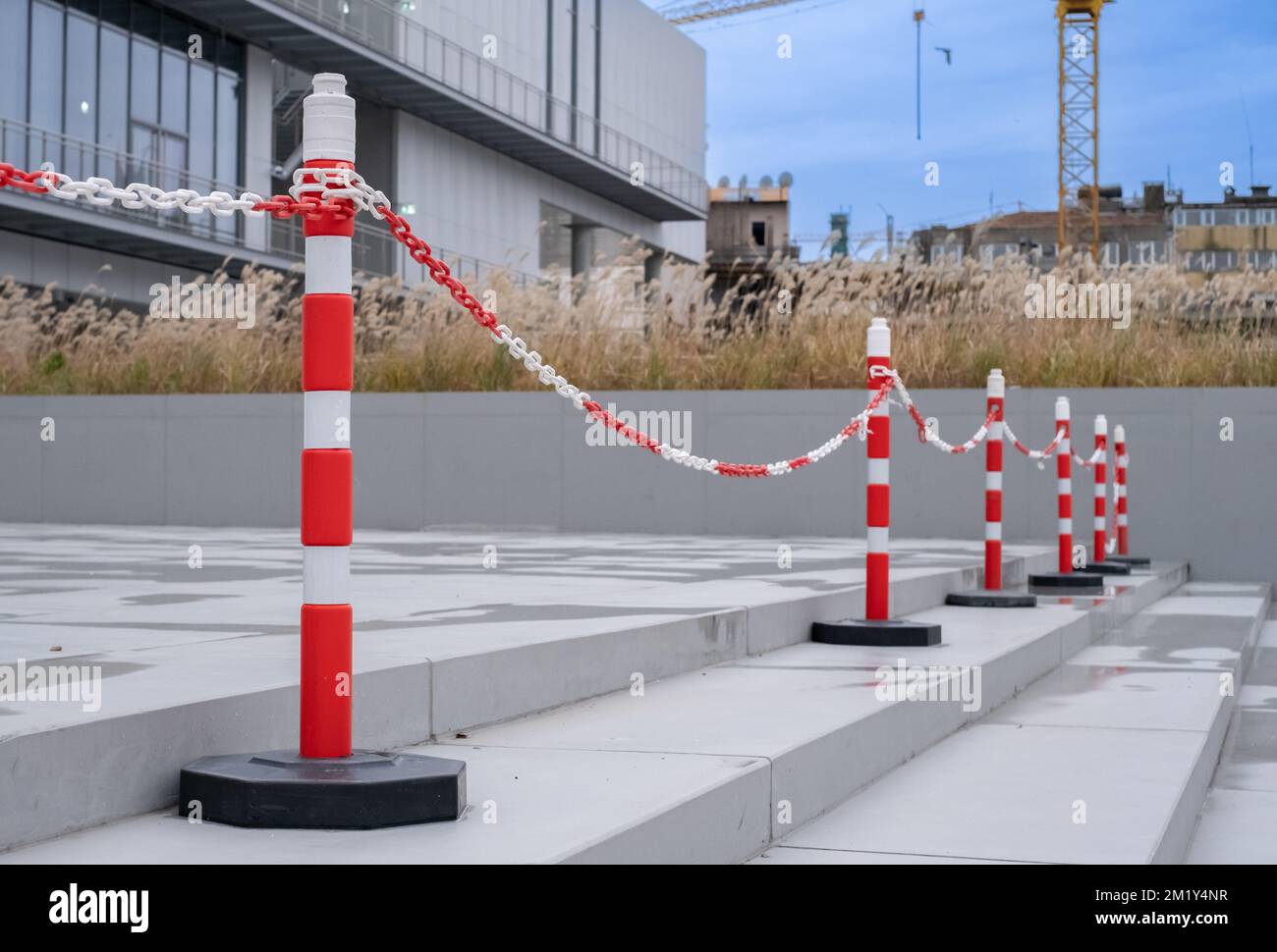 Side view of orange plastic street cones and poles with reflective silver tape near chain link fence at the entrance of a building. Stock Photo