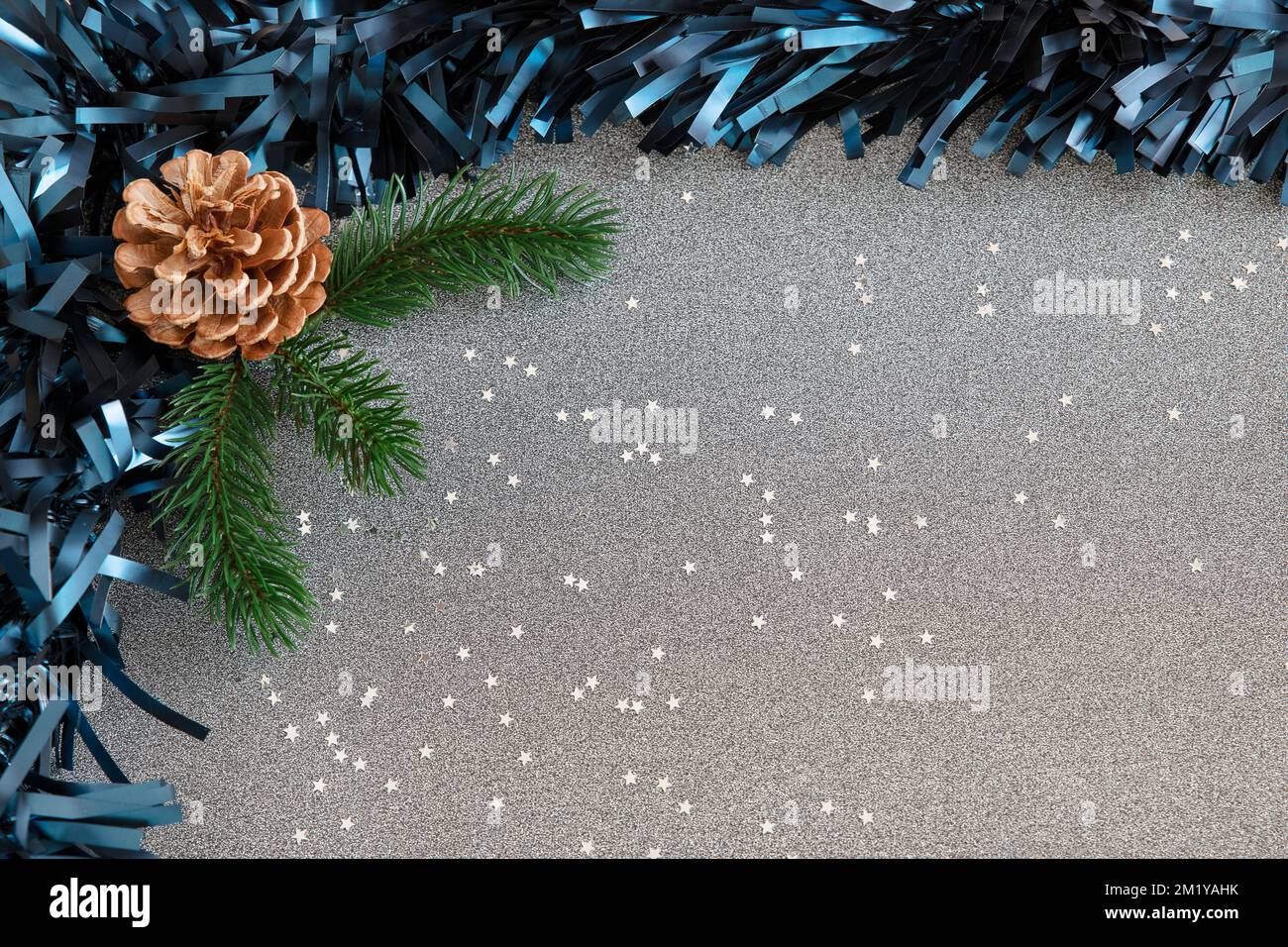Festive frame with blue tinsel, a pine cone and fresh pine brunches, and sparkling confetti stars spread against a silver gray background Stock Photo