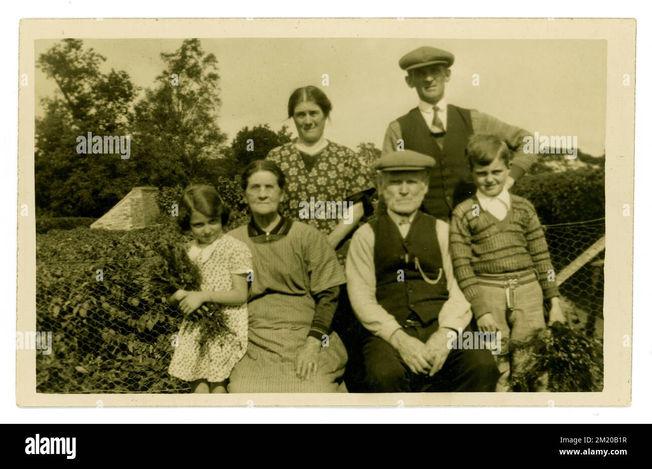 Original early 1930's era postcard of young children visiting their older rural farming relatives, grandparents or aunts and uncles on a British farm, growing vegetables. The men wear flat caps and suits and the women housecoats. U.K. Stock Photo