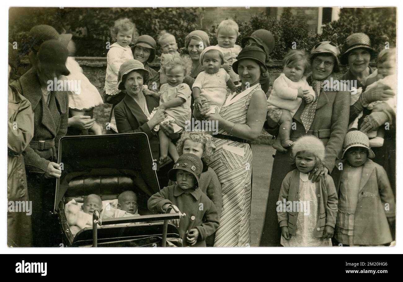 Original early 1930's era postcard of group of animated lively group of mothers outdoors on a Spring day with many small children, toddlers, preschool years, babies in arms, some are carried in their mothers arms, twins in a pram, Many characters. having lots of fun, smiling, maybe a mothers and babies group outing. Fashionable cloche hats on mums and girls.(fashionable until 1933) Coats with large collars, defined belted waist, date image to circa 1930 / 1931 U.K. Stock Photo