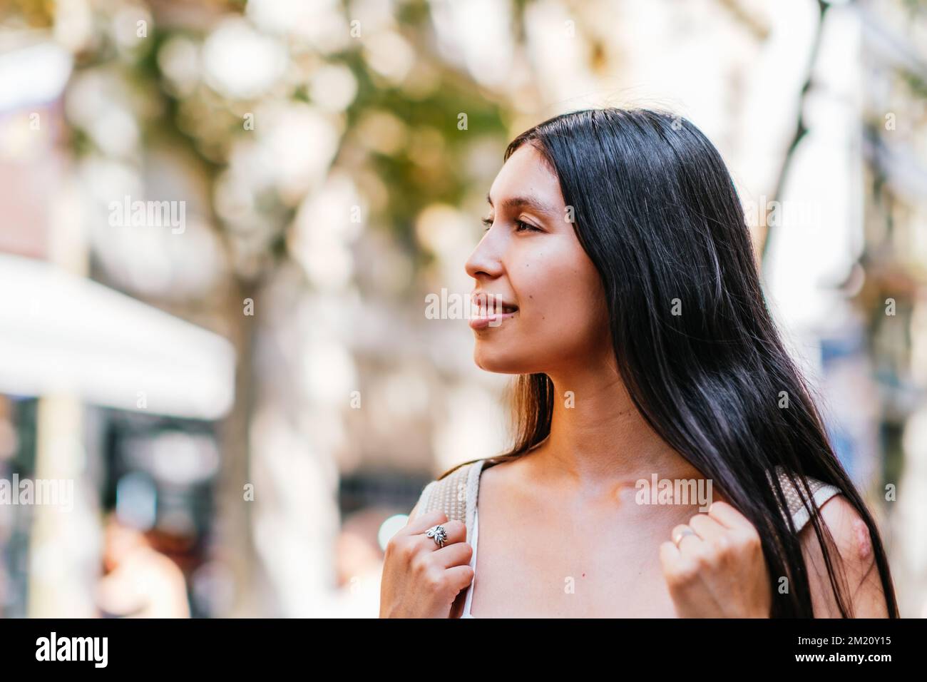 Positive Hispanic female tourist with long black hair looking away with smile on blurred background of sunlit street in Barcelona, Spain Stock Photo