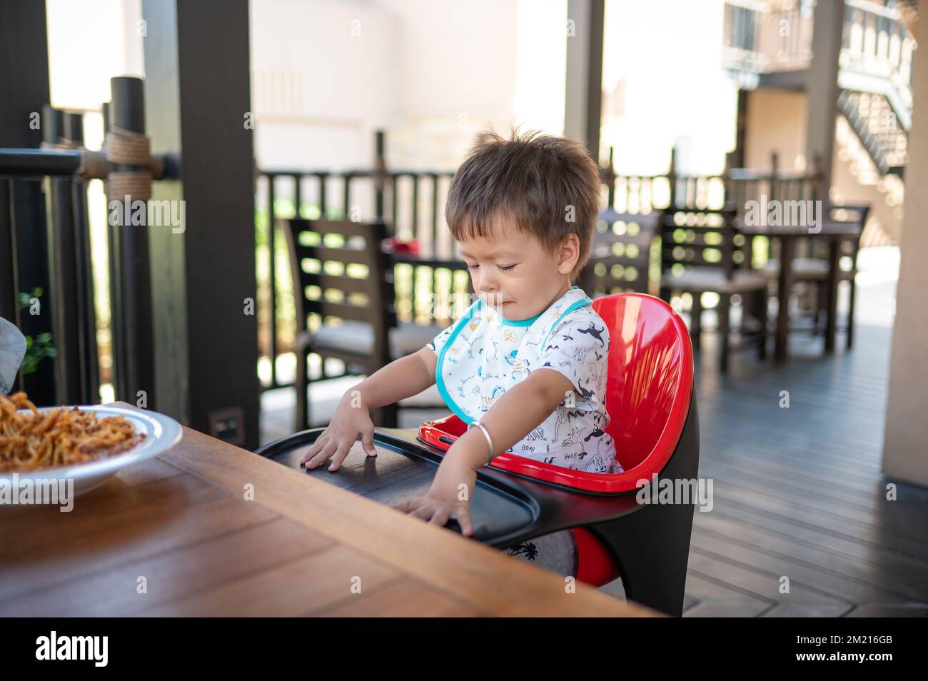 Toddler sitting on a high chair in the restaurant during a meal. Handsome multiracial one and half year old baby boy having a meal outdoors Stock Photo