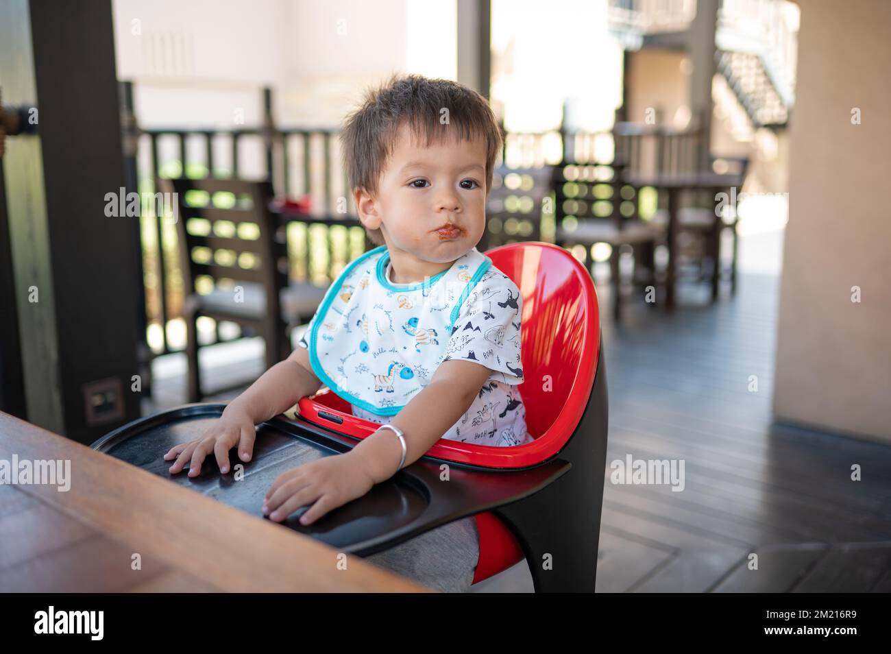 Toddler sitting on a high chair in the restaurant during a meal. Handsome multiracial one and half year old baby boy having a dinner meal in the resta Stock Photo