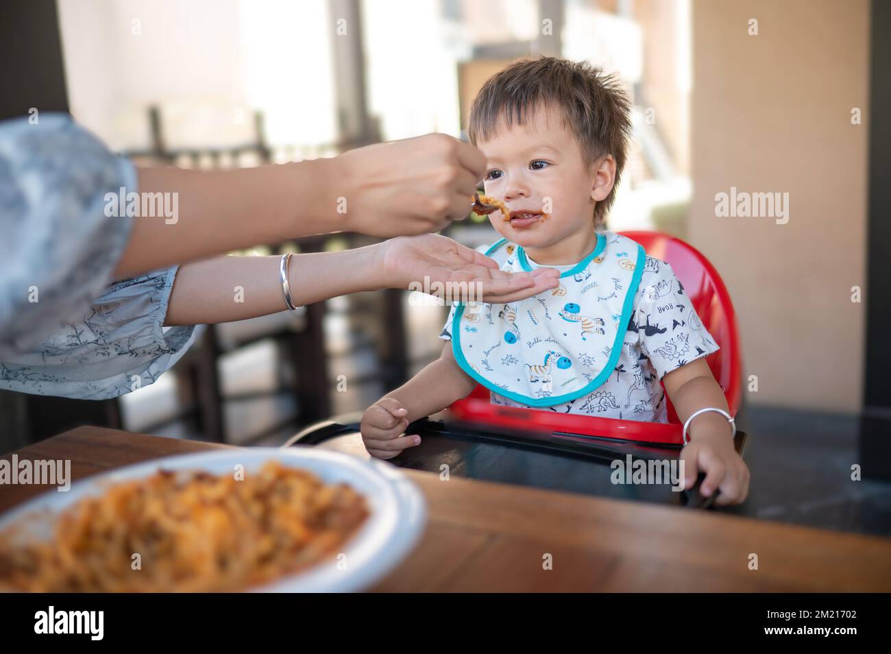 Toddler being fed in the restaurant by his mom. Handsome multiracial one and half year old baby boy having a dinner meal in the restaurant with his mo Stock Photo