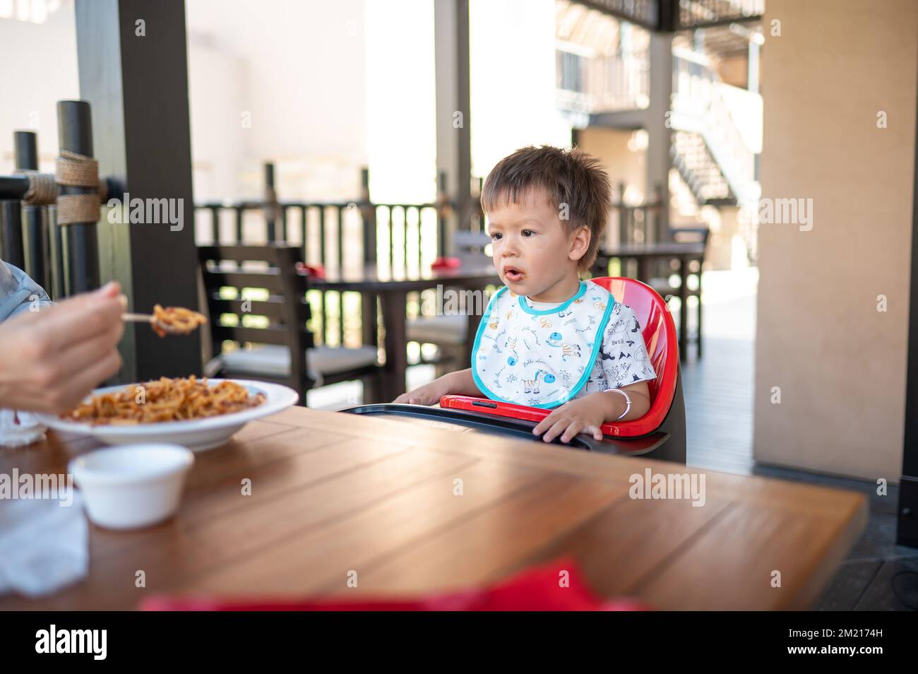 Toddler sitting on a high chair in the restaurant during a meal. Handsome multiracial one and half year old baby boy having a meal outdoors Stock Photo