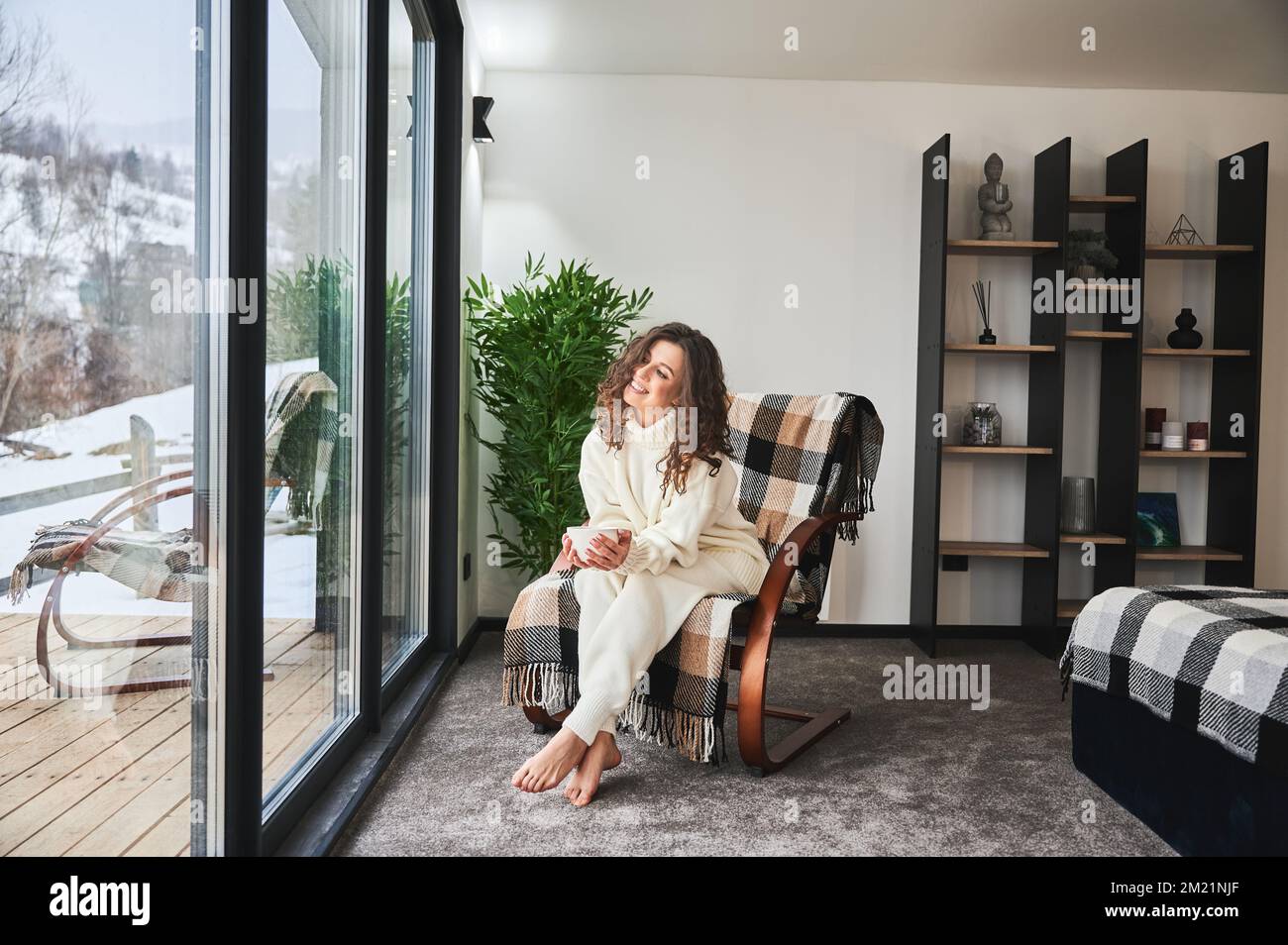 Curly young woman resting inside contemporary barn house in the mountains. Happy female tourist sitting on chair, holding cup of tea, enjoying view of mountain landscape through panoramic windows.. Stock Photo