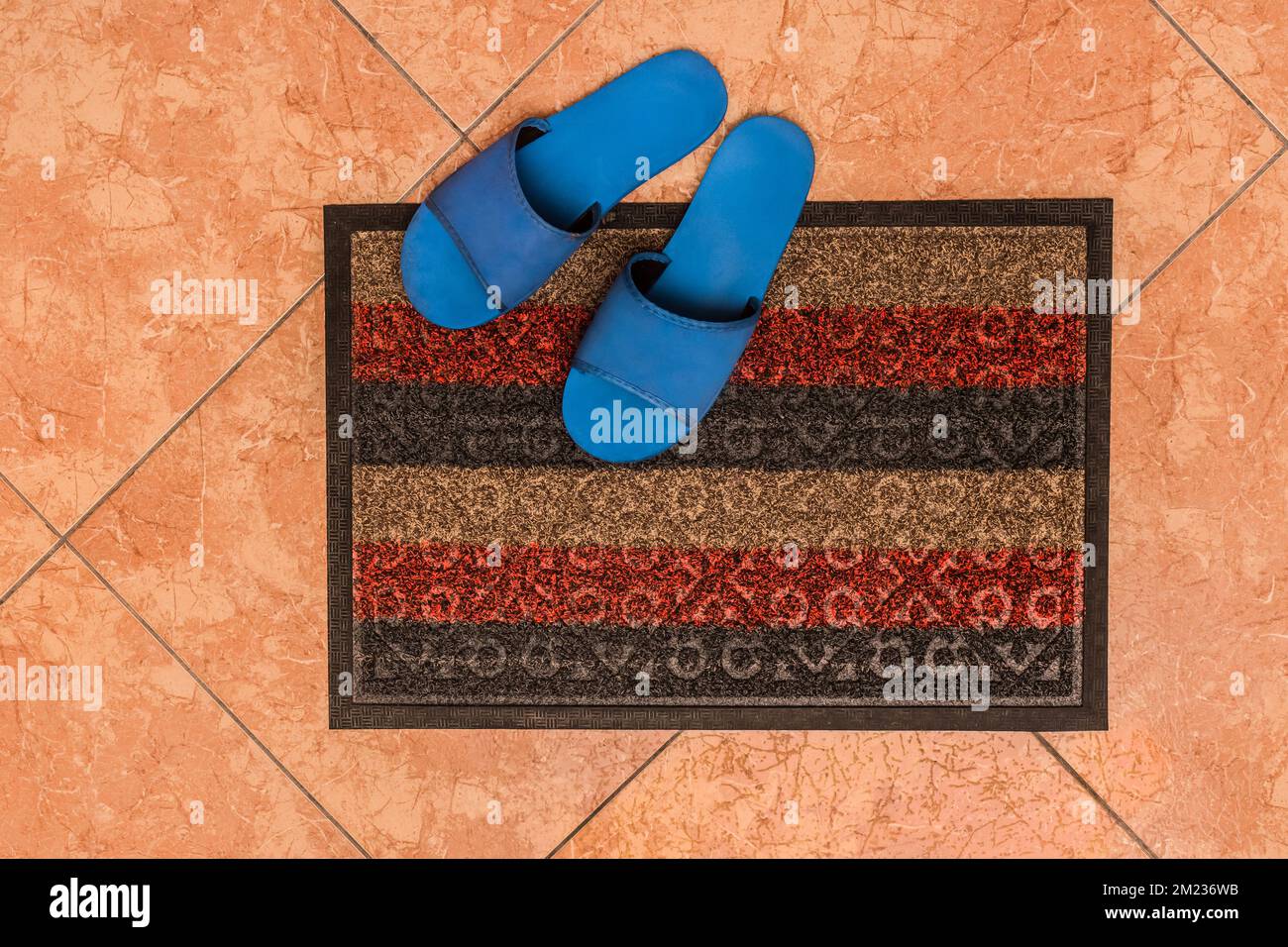 Blue homemade slippers stand on the foot carpet against the background of the floor tiles, the top view from above. Stock Photo