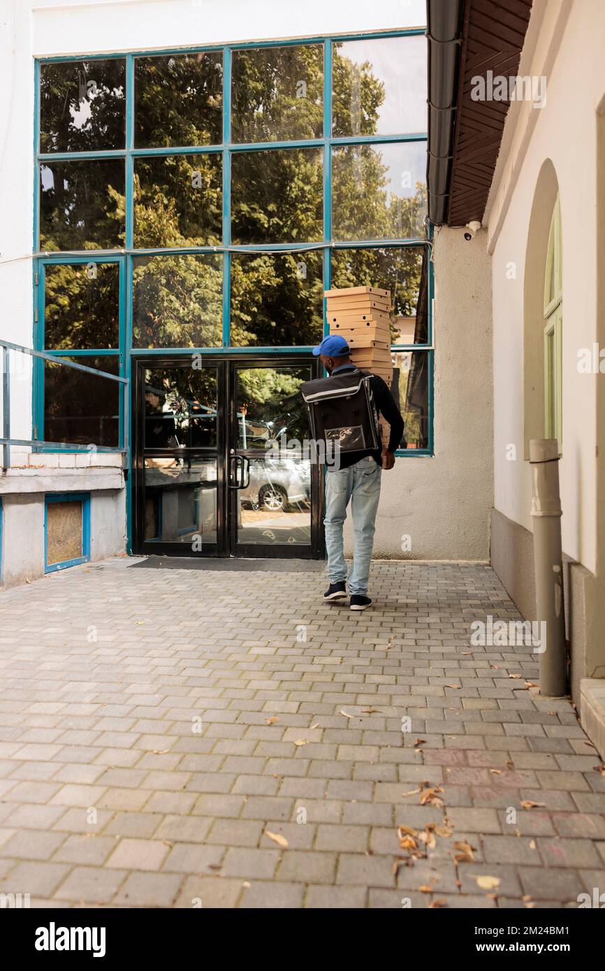 Deliveryman carrying huge pizza boxes pile to office back view, courier holding food order outdoors. Takeaway lunch delivery service african american worker delivering fastfood Stock Photo