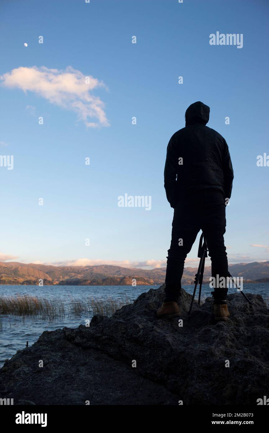 A young male photographer silhouette standing up over a rock with blue ...