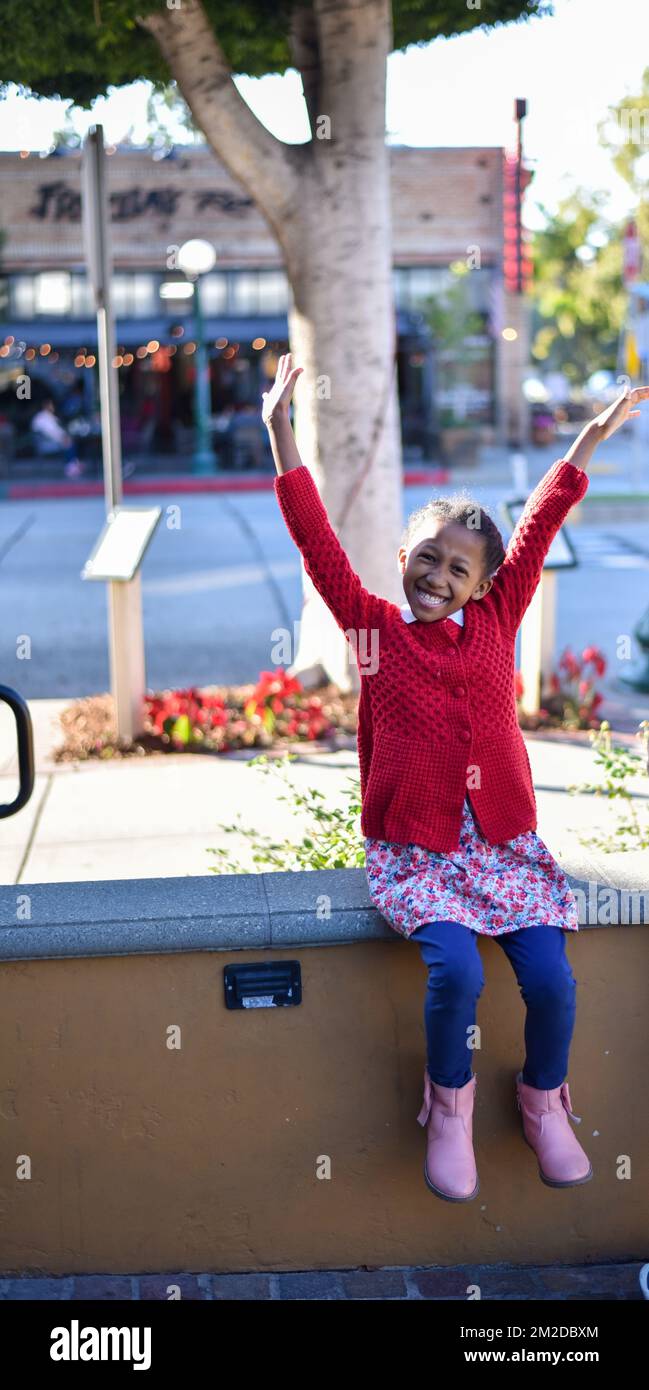 young girl wearing a bright red sweater expressing excitement and happiness with hands raised high in the air , smiling big Stock Photo