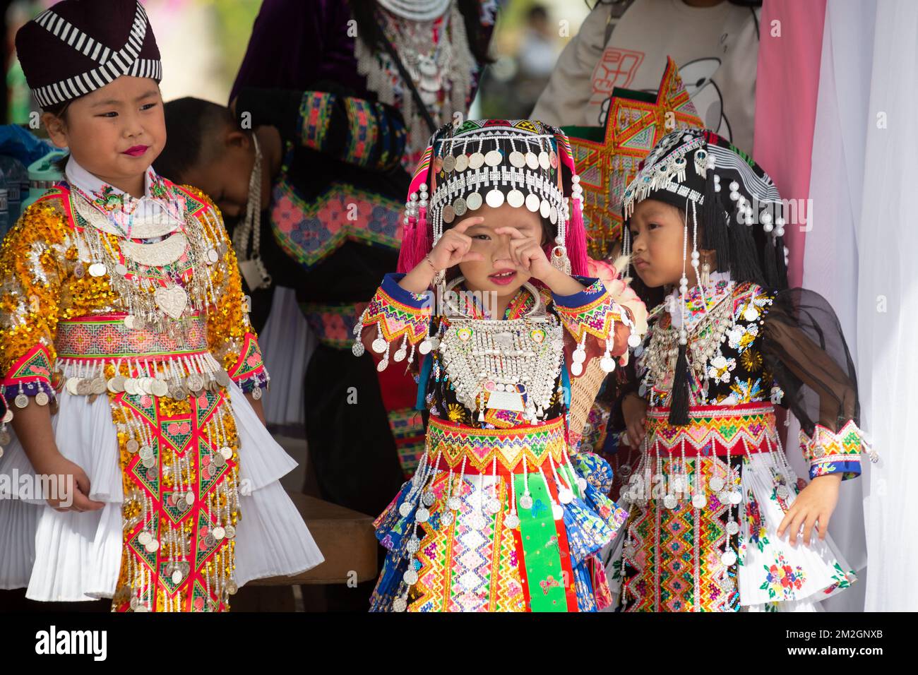 A young girl standing with friends in traditional Hmong garb retends to