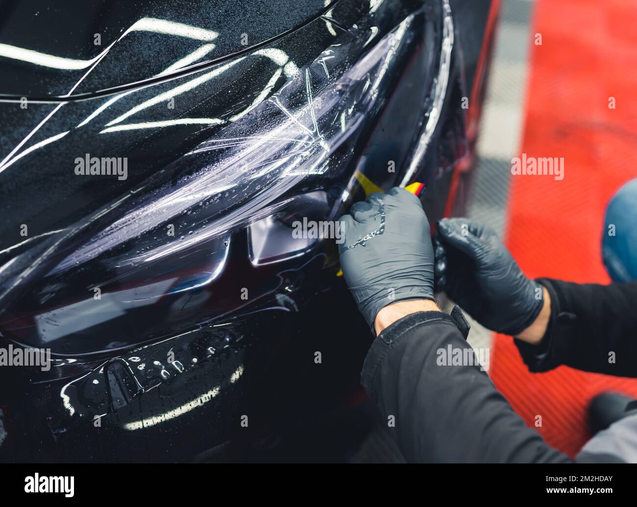 Two hands in protective black rubber gloves covering vehicle headlight with film to darken them. Closeup indoor shot. Car detailing. High quality photo Stock Photo