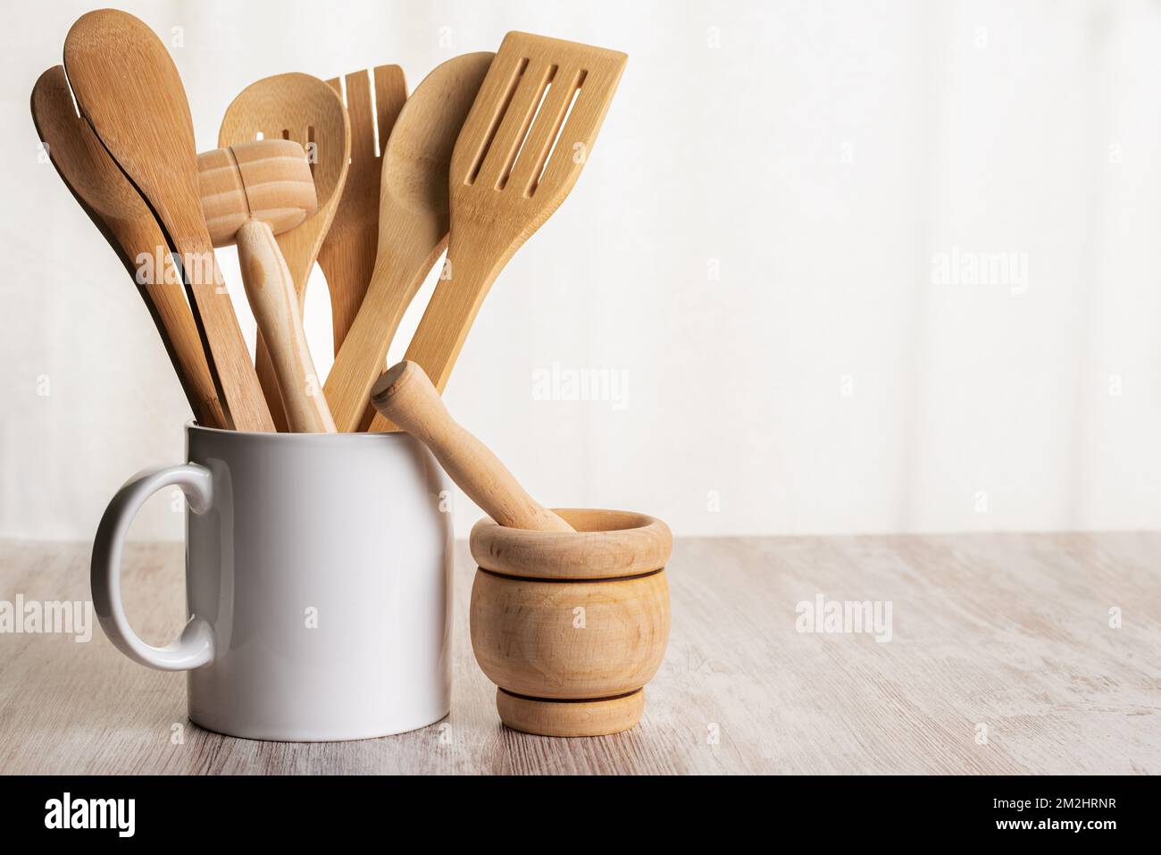 Wooden kitchen utensils such as spoons, forks and tongs inside a jar and next to a kitchen mortar on a wooden counter. Stock Photo