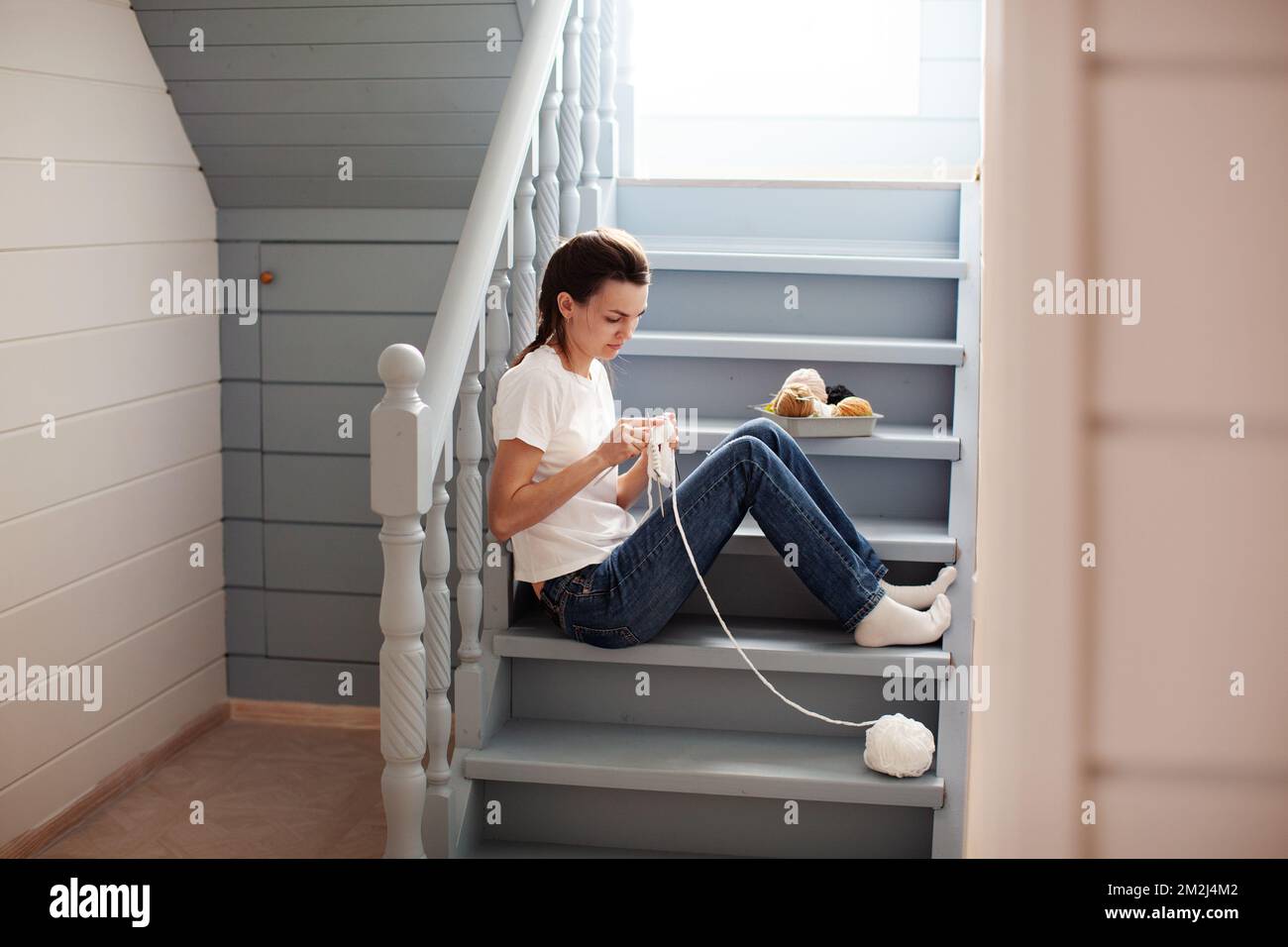 Calm woman knit white thread on circular knitting needles, sitting on steps in cozy wooden interior house. Crafting and handmade. Lifestyle and beauti Stock Photo