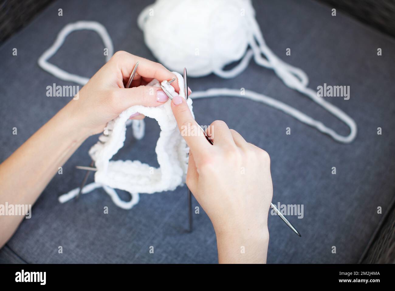 Woman hands knit white thread on circular knitting needles, dark blue blurred background. Crafting and handmade Stock Photo
