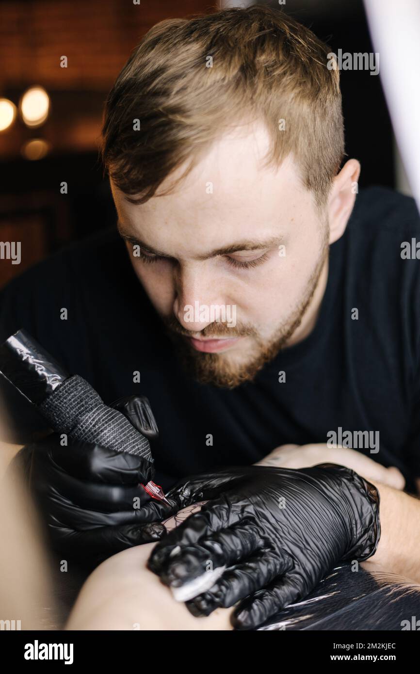 Close-up of a young professional tattoo artist introduces black ink into the skin using needle from a wireless tattoo machine. Tattoo on hand. Stock Photo